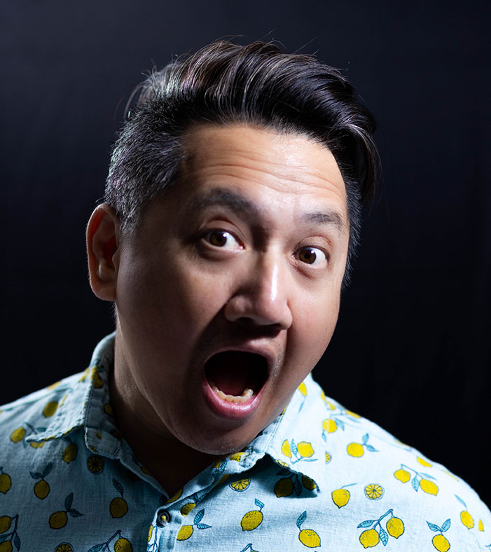 John Vuong Close-up portrait of a man with styled dark hair making a wide-eyed, open-mouthed surprised expression while wearing a light blue lemon-patterned shirt.